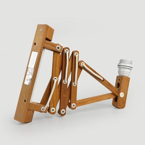 Peut inclure: Une applique murale en accordéon fabriquée en bois brun clair avec un cordon blanc passant à travers les articulations. La base comporte une étiquette rectangulaire blanche avec du texte noir et une douille blanche est fixée à l'extrémité.