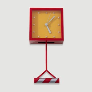 Op de afbeelding: Een rode en gele vierkante klok met zilveren wijzers en een rode standaard. De wijzerplaat is geel met kleine vierkante markeringen. De standaard heeft een rode paal en een basis met rode en zilveren strepen. Een moderne, decoratieve klok.