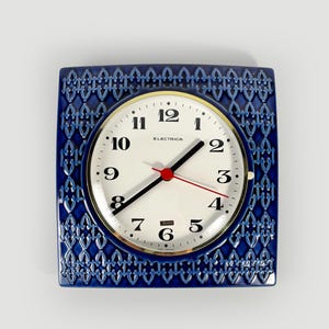 May include: A square wall clock with a deep blue ceramic frame featuring a repeating lattice pattern. The circular clock face is white with black Arabic numerals, black hour and minute hands, a red second hand, and the text Electrica.