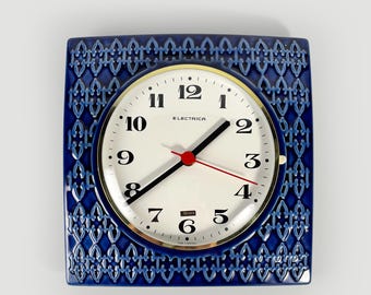 Puede incluir: Un reloj de pared cuadrado con un marco de cerámica azul profundo que presenta un patrón de celosía repetitivo. La esfera circular es blanca con números arábigos negros, manecillas negras, un segundero rojo y el texto Electrica.