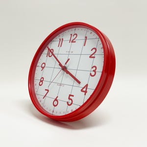 May include: A red quartz wall clock with a white face and red numerals. The clock has a grid pattern on the face and red hands. The clock is round and has a red bezel.