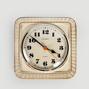 May include: A square wall clock with a cream-colored ceramic frame featuring a textured grid pattern. The round clock face is cream with black Arabic numerals, black hour and minute hands, and a thin orange second hand. Text reads DIPLOMAT and QUARTZ.