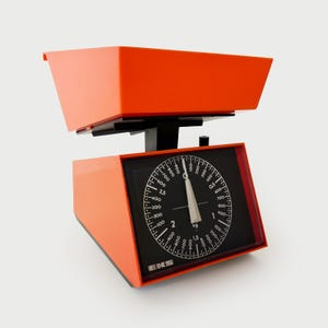Puede incluir: Una báscula de cocina naranja vintage con una esfera negra y marcas blancas. La báscula tiene una base rectangular y una bandeja. La esfera muestra medidas en kilogramos. La marca "EKS" es visible.