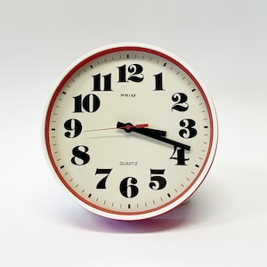 May include: A white analog clock with black numbers and hands, a red rim, and the words "PRIM" and "QUARTZ" printed on the face.