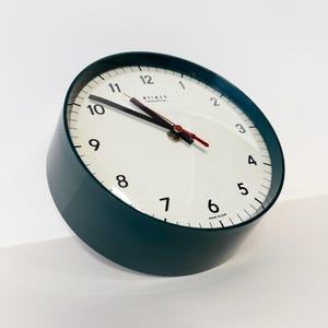 Puede incluir: Un reloj analógico de color verde azulado con una esfera blanca. El reloj tiene manecillas negras, un segundero rojo y números negros. La esfera del reloj tiene la marca "WEIMAR" impresa en la parte superior. El reloj está ligeramente inclinado.