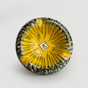 Puede incluir: Un cuenco de cerámica redondo con un centro amarillo brillante y rayas verde oscuro radiantes. El borde grueso y texturizado está moteado con tonos negros, blancos y verdes. Una pegatina cuadrada en el centro muestra el texto Hungaria y Zsolnay.