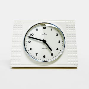 Może przedstawiać: Biały, prostokątny budzik z okrągłą tarczą. Tarcza zegara ma czarne cyfry i wskazówki, z widoczną marką "JUNGHANS electronic". Rama zegara ma teksturowany wzór po bokach.