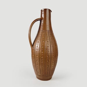 Può includere: Una brocca in ceramica marrone alta con un manico singolo e un collo stretto. Il corpo presenta scanalature verticali che si alternano a file di motivi in rilievo a forma di stella e cerchio, creando una superficie strutturata sull'esterno.