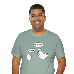 May include: A teal green t-shirt with a cartoon chicken and egg design. The chicken is saying "No me first!" and the egg is saying "Me first!"