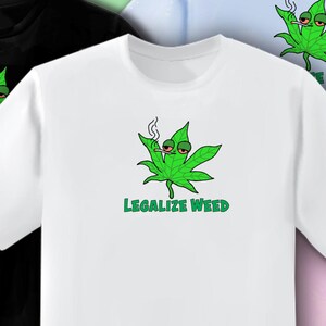 May include: White t-shirt featuring a green cannabis leaf character with red eyes and a lit cigarette. The text "LEGALIZE WEED" is printed in green below the graphic. The shirt is a crew neck style.