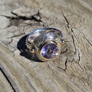 May include: A silver ring with a large, oval, light purple gemstone set in a gold bezel. The ring has a textured, ribbed design on the band and small gold accents on either side of the gemstone. The ring is displayed on a weathered wooden surface.