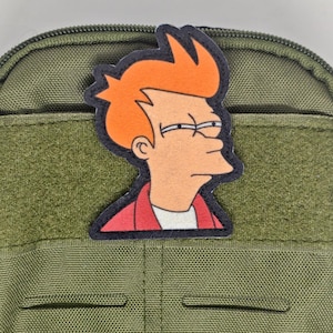 May include: An olive green tactical pouch featuring a rectangular hook-and-loop panel on the front. Attached to the panel is a colorful embroidered patch depicting a cartoon character with bright orange hair, squinting eyes, and a red jacket.