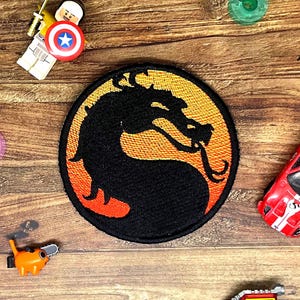 May include: A circular embroidered patch featuring a black dragon silhouette centred against a vibrant orange and yellow gradient background. The patch has a thick black stitched border and is surrounded by various small plastic toy figures.