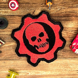 May include: A circular embroidered patch featuring a red gear shape with a black centre and a red skull graphic in the middle. The patch has a thick black border and is displayed alongside various small toy items on a wooden surface.