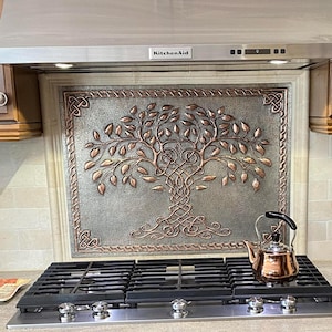 May include: A rectangular metal wall plaque featuring a raised copper-coloured tree of life design with intricate Celtic knotwork branches and roots, mounted as a decorative backsplash above a stainless steel gas hob and cooker hood.
