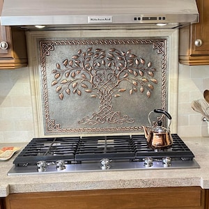 May include: A kitchen with wooden cabinets, a stainless steel range hood, and a gas cooktop. A copper tea kettle sits on the cooktop. A decorative backsplash features a tree design. A coffee maker and utensils are on the countertop.