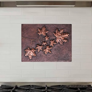 Puede incluir: Un protector contra salpicaduras de metal de color cobre con un diseño en relieve de una rama de hoja de arce. El protector contra salpicaduras está instalado sobre una estufa de acero inoxidable con cinco perillas de quemador.