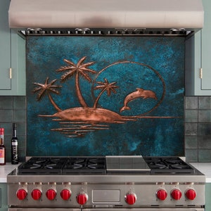 Op de afbeelding: Een decoratief metalen achterwandpaneel met een koperkleurig reliëf van een tropisch eiland met palmbomen en een springende dolfijn tegen een getextureerde, donkerblauwe achtergrond, geïnstalleerd achter een roestvrijstalen fornuis.