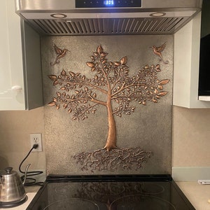 May include: A rectangular metal backsplash panel featuring a raised copper-toned relief of a tree with exposed roots and two hummingbirds. The background has a textured, metallic gray finish. The panel is installed behind a black glass stovetop.