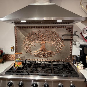 May include: A rectangular copper-toned metal backsplash panel featuring a central embossed tree of life design with intricate knotwork borders, installed behind a stainless steel gas stove with a black floral patterned tea kettle on top.
