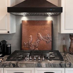 May include: A black range hood with a copper backsplash featuring two birds perched on a branch with flowers. The backsplash is a decorative metal panel with an embossed design.