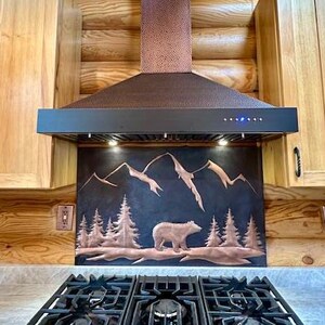 May include: A copper range hood with a hammered texture sits above a stove. Below the hood, a metal backsplash features a relief design of a bear walking through a forest with mountains in the background. The hood has a black control panel.