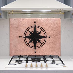 May include: A copper-colored backsplash with a black compass design above a white gas stove. The compass rose is centered, with the cardinal directions labeled. The stove has black grates and gold-colored knobs. The range hood is white.