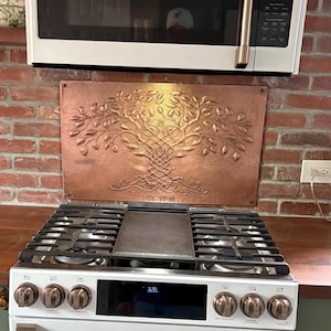 May include: A rectangular copper backsplash panel featuring a raised, embossed tree of life design with intricate Celtic knotwork roots and branches. The metal surface has a hammered texture and includes the engraved text EST. 1949 at the bottom.