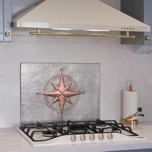 Puede incluir: Una cocina con una campana extractora blanca y dorada sobre una placa de gas. Una placa para salpicaduras decorativa de metal con un diseño de rosa de los vientos de cobre está centrada sobre la placa. Un portarrollos de papel con base dorada está a la derecha.