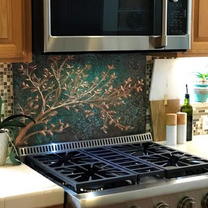 May include: A rectangular kitchen backsplash panel featuring a raised copper-coloured tree branch design against a textured dark teal and black background, positioned vertically between a stainless steel gas range and a microwave oven.