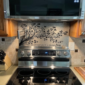 May include: A rectangular metal stove backsplash featuring a black tree branch silhouette and the text "The secret ingredient is always Love" in black script. The panel is installed behind a stainless steel electric range with a pot filler faucet.