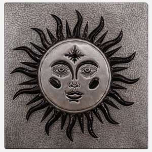 Puede incluir: Un sol plateado con una cara y una estrella en la frente, en relieve sobre un fondo gris.