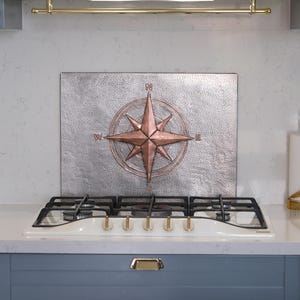 Puede incluir: Un panel protector de metal rectangular con un diseño de rosa de los vientos central en color cobre y los puntos cardinales N, S, E y O. El panel tiene un acabado plateado martillado y texturizado detrás de una estufa de gas blanca.