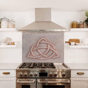 May include: A stainless steel range hood with a copper-colored Celtic knot design backsplash. The backsplash is a decorative accent for a kitchen.