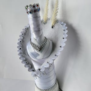 May include: A decorative bottle wrapped in white satin ribbon, adorned with a jeweled crown, necklace, and trim. The design includes a white felt heart-shaped collar with silver trim and decorative accents. The bottle is topped with two sprigs of dried grass.