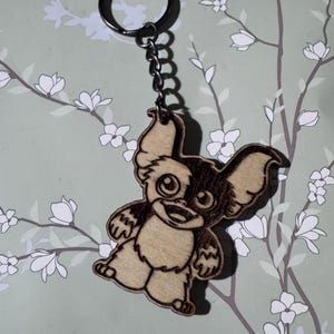 May include: A wooden keychain featuring a laser-engraved illustration of a small, furry creature with large ears, wide eyes, and a smiling mouth. The design is rendered in dark brown tones on a light tan wood surface, attached to a metal ring.