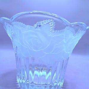 Peut inclure: Vase en verre transparent avec une anse et un motif floral de rose. Le vase présente une texture côtelée sur la partie inférieure et un bord festonné. Le motif de la rose est givré, créant un contraste avec le verre clair.