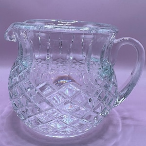 Op de afbeelding: Transparante glazen kan met deksel, met een diamantgeslepen patroon. De kan heeft een gebogen handvat en een schenktuit. Het ontwerp is klassiek en elegant, geschikt voor het serveren van dranken.