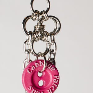 May include: A silver-toned metal earring featuring a chainmail-style link design connected to a round, hot pink plastic button. The button is embossed with the white text Cabbage Patch Kids and has two central holes, suspended by silver metal chains.