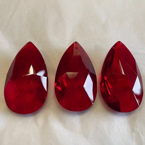 May include: Three faceted, teardrop-shaped red glass pendants. Each pendant has a pointed top and a rounded bottom. The pendants are a deep, rich red colour and appear to be made of a transparent material. They are arranged in a row.