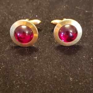 May include: A pair of gold-toned cufflinks with a round, deep red center. The cufflinks have a classic design, with a polished finish. The background is a dark, textured surface.