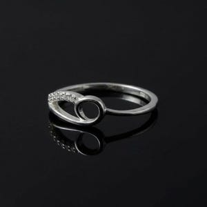 May include: A silver ring with a distinctive design. The ring has a curved form with a small circle and a section embellished with tiny, clear stones. The ring is shown against a dark backdrop, accentuating its reflective surface.