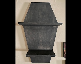 May include: A dark gray wooden shelf with a unique coffin-like shape. The shelf has two horizontal ledges for displaying items. The wood grain is visible, adding texture to the design. It is mounted on a light-colored wall.