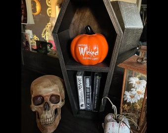 May include: A black wooden coffin-shaped shelf with a pumpkin on the top shelf and five books on the bottom shelf. The pumpkin is orange and has the words "Something Wicked This Way Comes" written on it. The books are all black with white text.