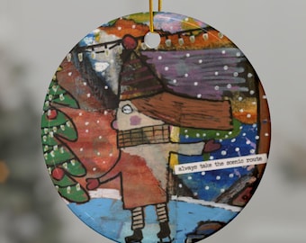 May include: A round Christmas ornament with a whimsical illustration of a person ice skating near a Christmas tree. The ornament has a gold string for hanging and the text "always take the scenic route".