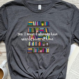 May include: A dark gray t-shirt with a graphic of a bookshelf full of colorful books and plants. The text "yes, I know I already have unread books at home" is printed on the shirt.