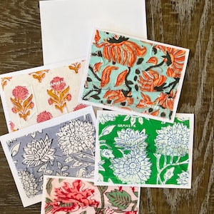 May include: A collection of square greeting cards with floral patterns in various colors, including orange, pink, blue, and green. Each card has a white border and the word "NAMASTE" printed on it. The cards are arranged on a wooden surface.