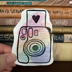 May include: A colorful sticker of an instant camera with a watercolor effect. The camera has a rainbow lens and the sticker has handwritten text that reads "I want to see the world with my own eyes, all of it. The beauty..."
