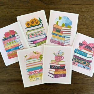 May include: A collection of six greeting cards with illustrations of stacked books. Each card features a different floral arrangement atop the books, including sunflowers, roses, and hydrangeas. The cards have a pink border and are arranged on a wooden surface.