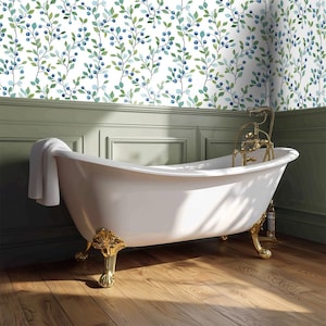 May include: A claw-foot bathtub with gold-colored accents and fixtures is the focal point of this bathroom. The white tub is set against a backdrop of green wainscoting and a wallpaper pattern of blue berries and green leaves. A white towel hangs on the tub.